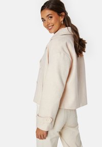 Světle růžová, zkrácená bunda z vlny se šálovým límcem, dvouřadovým zapínáním a postranními kapsami; spárovaná se světle béžovými kalhotami.