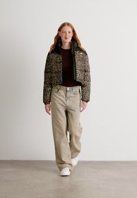 Pelsprøve av leopard med kort pufferjakke med svart trim, paret med løse beige bukser og hvite sneakers, satt mot en ensfarget bakgrunn.