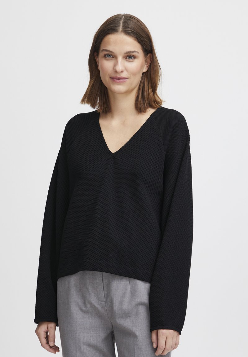 Pull noir texturé avec un col en V et de longues manches larges, associé à un pantalon gris clair. Le pull a une coupe décontractée et des détails en côtes.