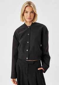 Musta lyhyt jakku, jossa on pyöreä pääntie, harmaat napit ja pitk�ät hihat. Öljymäinen kangas, jossa on hieman kiiltoa ja sivutaskut.