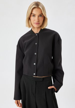 Black cropped jacket with a round neckline, featuring grey buttons and long sleeves. Smooth fabric with a slight sheen and side pockets.