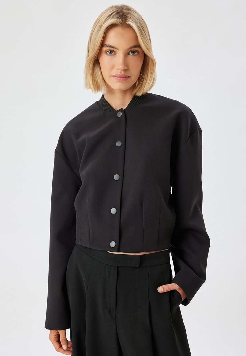 Musta lyhyt jakku, jossa on pyöreä pääntie, harmaat napit ja pitkät hihat. Öljymäinen kangas, jossa on hieman kiiltoa ja sivutaskut.