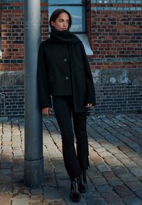 Cappotto di lana nera con un colletto alto e due bottoni, abbinato a pantaloni neri attillati e stivaletti, sullo sfondo di un muro di mattoni e ciottoli.