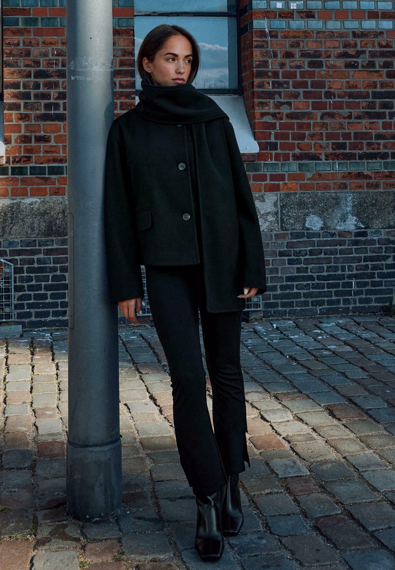 Cappotto di lana nera con un colletto alto e due bottoni, abbinato a pantaloni neri attillati e stivaletti, sullo sfondo di un muro di mattoni e ciottoli.