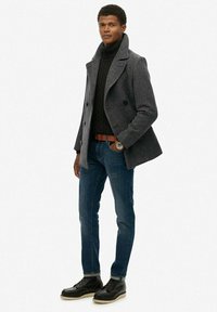 Abrigo de lana gris de doble botonadura sobre un cuello de tortuga de punto negro, jeans ajustados azules y botas negras con suelas beige. Reloj plateado como acento.