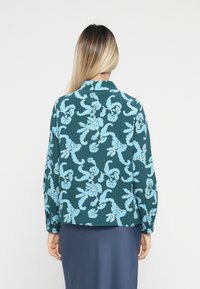 Teal bluse med blomsterdesign i lys blå, med lange ermer, knaplukning ved håndleddene og en lige kant. Synlig tekstur og kragedetaljer.