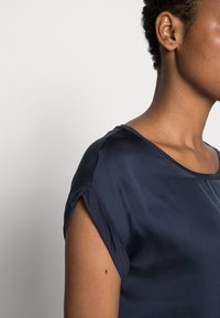 Top blu navy a maniche corte con scollatura rotonda, texture morbida e maniche arrotolate, caratterizzato da una vestibilità rilassata e un tessuto liscio.