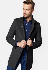 Węgorzowy płaszcz w strukturę jodełki, zapinany na jeden guzik, z wyłogowym kołnierzem, noszony na ciemnoniebieskiej koszuli i w kratkę spodni.