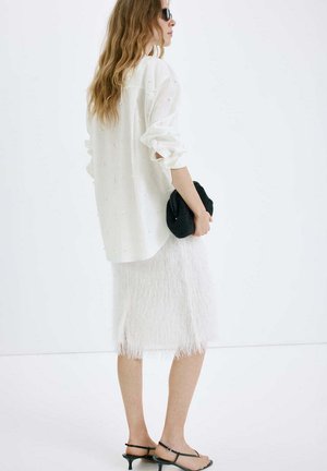 Mujer con blusa blanca de manga larga y falda midi blanca texturizada, sosteniendo un bolso de mano negro y llevando sandalias negras con tacón.