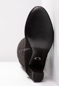 Botín de ante negro con un pequeño tacón, cremallera lateral y suela de goma texturizada. Base plana con un logo circular en el tacón.
