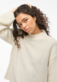 Beežist kootud kampsun kõrge ribamustriga kaelusega, liialdatud lõikega ja kukkunud õlgadega, millel on ühtlane tekstuuriline muster.