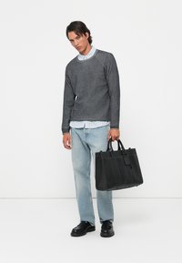 Grau strukturierter Pullover, hellblau gestreiftes Hemd, hellblaue Jeans, schwarze Lederhandtasche, schwarze Schuhe, stehend vor einem weißen Hintergrund.
