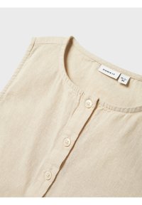 Chemise beige sans manches avec un col arrondi et une patte de boutonnage à cinq boutons. Fabriquée en tissu léger à la texture douce. Étiquette visible à l'intérieur.