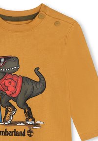 Oranje katoenen T-shirt met korte mouwen, voorzien van een grafiek van een dinosaurus die een zonnebril en laarzen draagt, en accentueert met knopen op de schouder.
