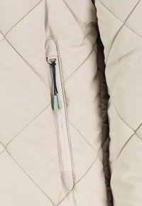 Tissu matelassé beige avec une poche zippée verticale comportant des cordons noirs et des languettes en plastique vert clair.
