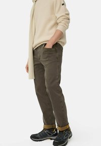 Persoon in opgerolde donkerbruine broek, lichtbeige geribde sweater en sjaal, met zwart en gele wandelschoenen, staand met één hand in de zak.