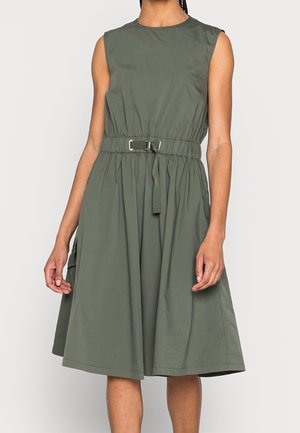 Robe sans manches vert olive longueur genou avec taille cintrée et ceinture à boucle portée par une personne debout sur un fond uni.