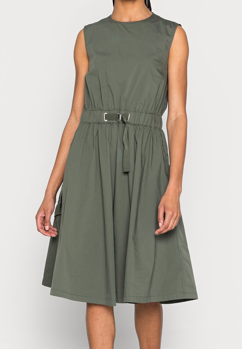 Robe sans manches vert olive longueur genou avec taille cintrée et ceinture à boucle portée par une personne debout sur un fond uni.