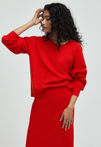 Roter Strickpullover mit langen, elastischen Ärmeln und lockerer Passform, kombiniert mit einem passenden roten Rock, der eine strukturierte Oberfläche zeigt.