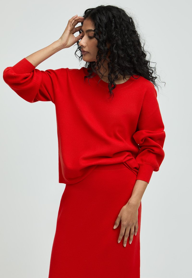 Roter Strickpullover mit langen, elastischen Ärmeln und lockerer Passform, kombiniert mit einem passenden roten Rock, der eine strukturierte Oberfläche zeigt.