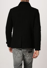 Giacca nera in misto lana con colletto a punte e orlo dritto, con cuciture laterali e un design minimalista. Indossata sopra dei jeans grigi.