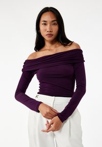 Lila off-the-shoulder toppi pitkillä hihoilla, jossa on rypytetty kangasviimeistely, yhdistettynä valkoisiin korkeavyötäröisiin housuihin ja minimalistisiin varusteisiin.