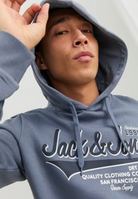 Niebieski sweatshirt z kapturem i sznurkiem, z dużym białym logo marki z przodu oraz miękką teksturą. Hoodie ma luźny krój.