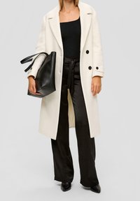 Abrigo texturizado en color ecru con botones negros, usado sobre una blusa negra acanalada y pantalones satén negros de pierna ancha, combinado con un bolso tote negro.