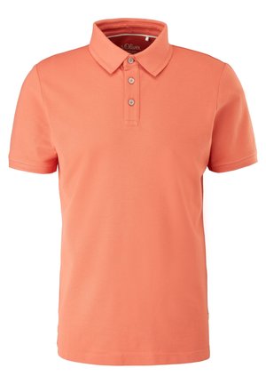 Homme portant un polo corail à manches courtes et un jean bleu clair, debout dans une posture détendue devant un fond uni.