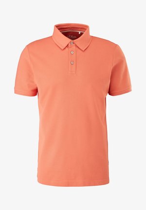 Mann trägt ein korallenfarbenes kurzärmeliges Polohemd und hellblaue Jeans, steht entspannt vor einem schlichten Hintergrund.