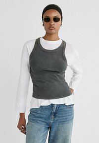 Canottiera grigia a coste indossata sopra una maglietta bianca a maniche lunghe. La canottiera presenta un collo rotondo e un design aderente. I jeans sono di un blu chiaro.