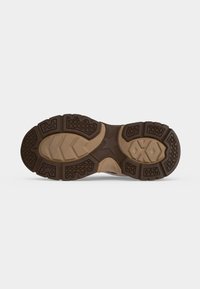 Semelle d'une chaussure robuste marron et beige avec des motifs de semelle texturés pour l'adhérence, montrée sur un fond clair uni.