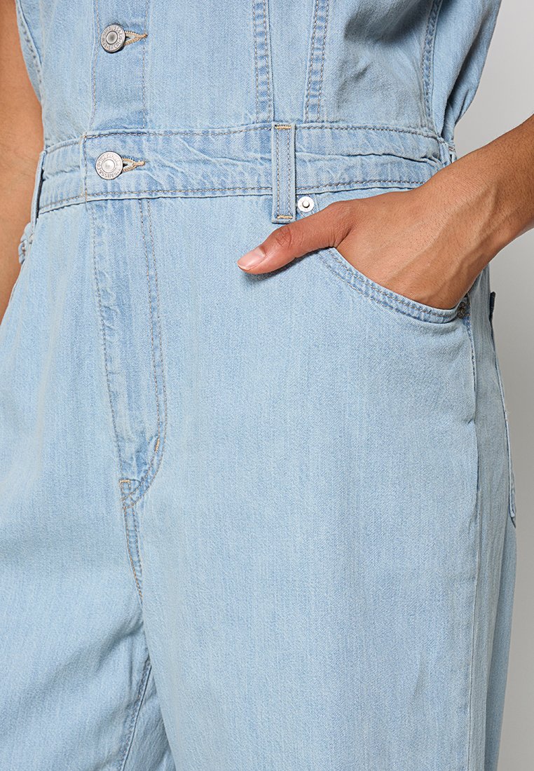 Salopette en denim bleu clair avec une taille cintrée, deux boutons en haut et des poches latérales. Texture douce et design à jambe droite.