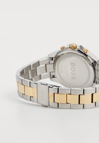 Montre pour homme avec un bracelet en acier inoxydable argenté et des accents dorés. Boîtier rond avec des boutons dorés et argentés, cadran blanc et logo au dos.