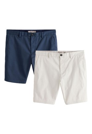 Ung mand står op ad en hvid væg iført en hvid t-shirt, marineblå shorts, hvide sokker og beige sneakers med et neutralt ansigtsudtryk.