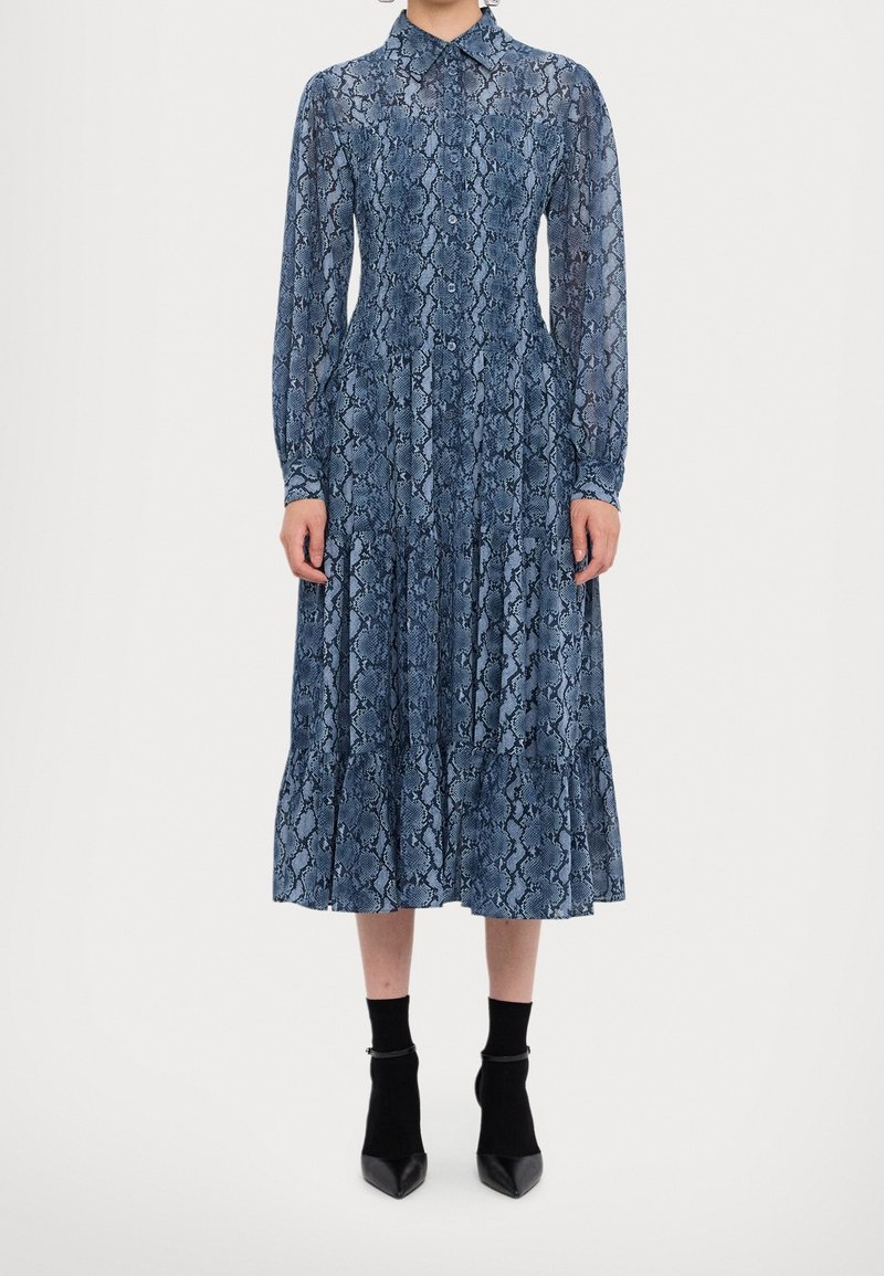 Femme portant une robe midi bleue à manches longues avec imprimé serpent, boutonnée, associée à des bottines noires et des chaussettes, debout droite sur fond blanc.