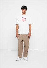 Camiseta de algodón blanca con el logo rojo de "Calvin Klein", combinada con pantalones beige. El modelo está de pie sobre un fondo liso. Zapatillas blancas completan el look.