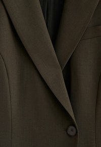 Veste cintrée vert olive foncé avec revers crantés, un bouton noir unique et un tissu texturé. Présente une coupe slim et une poche avant.
