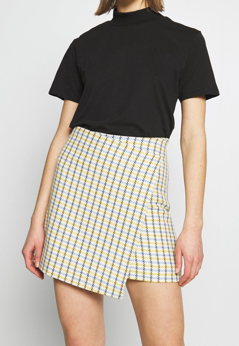 Femme portant un haut noir à manches courtes rentré dans une mini-jupe à carreaux jaune, bleu et blanc avec un ourlet asymétrique.