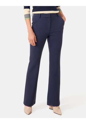 Femme portant un pantalon droit bleu marine, des chaussures beiges à bout pointu et un haut à manches longues avec des manches rayées et en blocs de couleur crème.