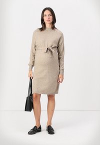 Abito a maglia beige con maniche lunghe, vita cinte da una cintura, e polsini a coste; abbinato a una borsa nera e mocassini neri.