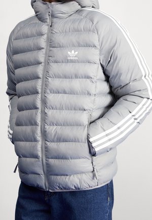 Man draagt een lichtgrijze gewatteerde Adidas-jas met witte drie strepen op de mouwen en handen in de zakken, gecombineerd met een blauwe spijkerbroek.