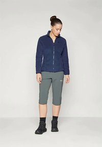 Marine fleecejakke med hel glidelås, kombinert med knelengde grå shorts. Teksturert stoff og glidelåsdetaljer synlige. Sorte sko fullfører antrekket.