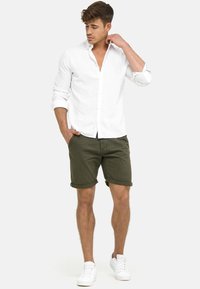 Hvid skjorte med knapper lavet af linned, parret med olivengrønne shorts oppe rullet. Modellen har hvide sneakers på og poserer med den ene hånd på kraven.