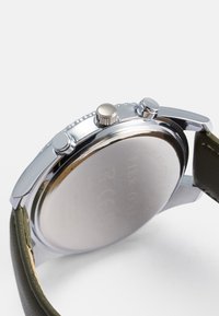 Nerūsējošā tērauda pulkstenis ar pulētu sudraba korpusu, zaļu ādas siksniņu un diviem spiedpogām. Iezīmes ietver gludu apaļu seju un aizmuguri ar gravējumu.