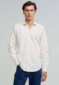 Off-white Hemd mit klassischem Kragen, langen Ärmeln und lockerer Passform, kombiniert mit dunkelblauen Jeans. Hat eine dezente Textur.