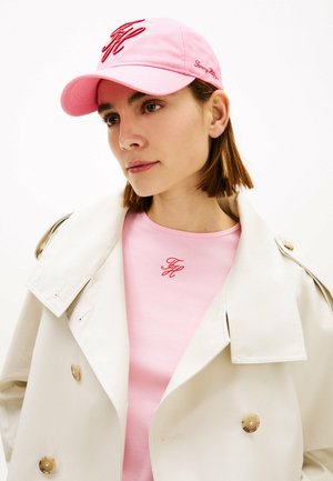Mujer con cabello corto y castaño, lleva una gorra rosa, camisa rosa con un logo rojo y un abrigo beige sobre un fondo blanco.