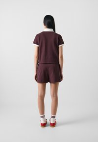 Burgundi színű, két részes együttes rövid ujjakkal és nadrágshorts-al, fehér galléros részlettel. Piros cipővel és csíkos zoknival párosítva.