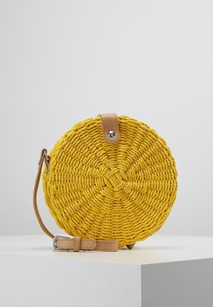 Sac à main rond jaune tissé avec une bandoulière en cuir beige et une fermeture à pression, présenté sur un socle blanc sur fond gris.