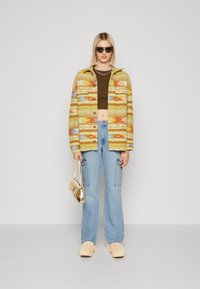 Kleurrijke jasje met patronen in geel, groen en rood, gecombineerd met een bruine crop top en lichtblauwe cargo jeans, accessorized met een gouden handtas.